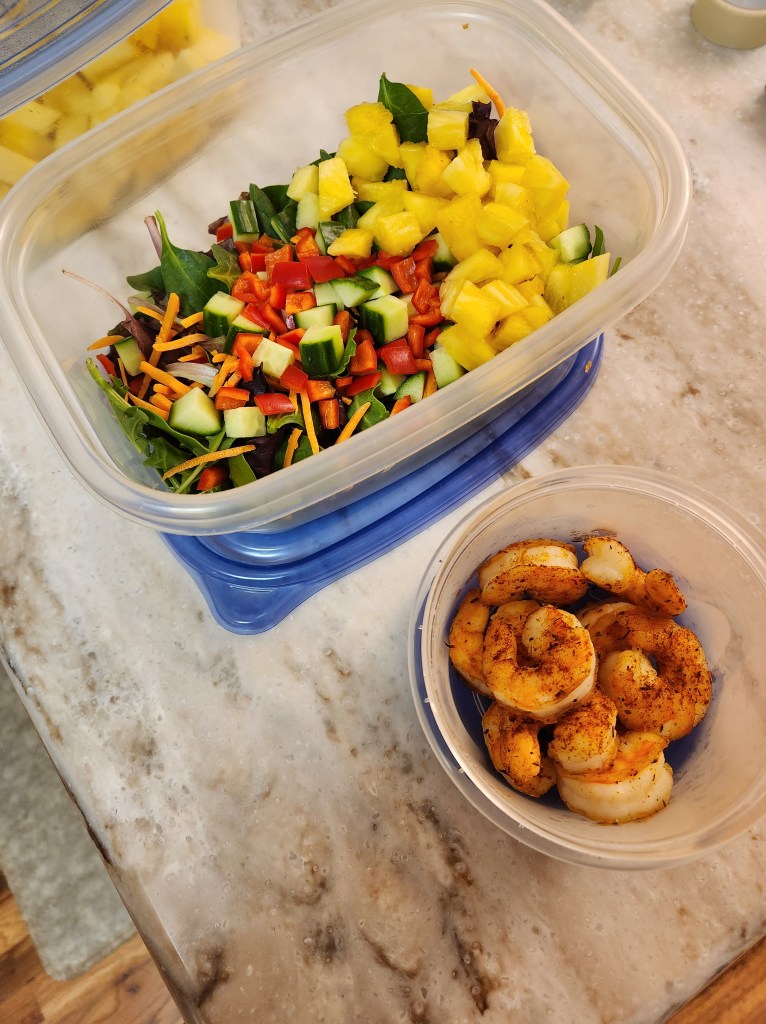 A power bowl prepped in a take away container with greens, chopped peppers, mangoes, cucumbers, and grilled shrimp.