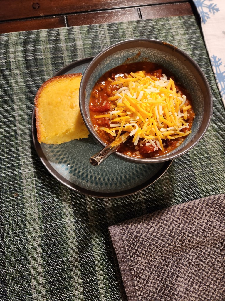 A scrumptious bowl of chili topped with cheese and a piece of homeade cornbread on the side.