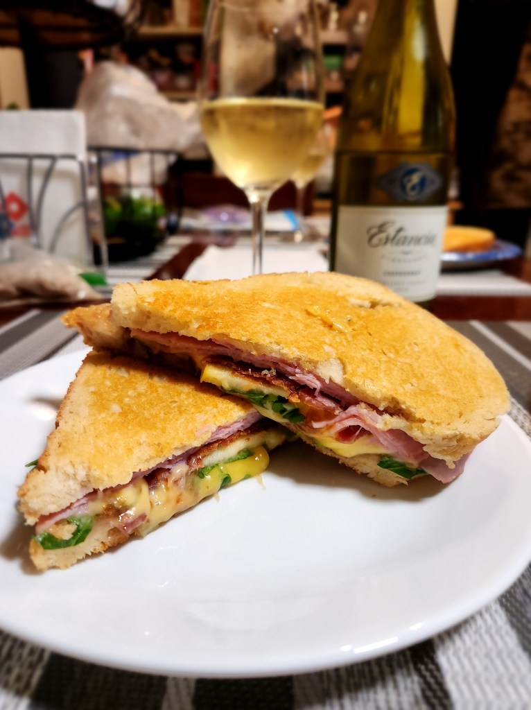 A parmesan crusted ham, turkey, and avocado sandwich with a glass of white wine in the background.