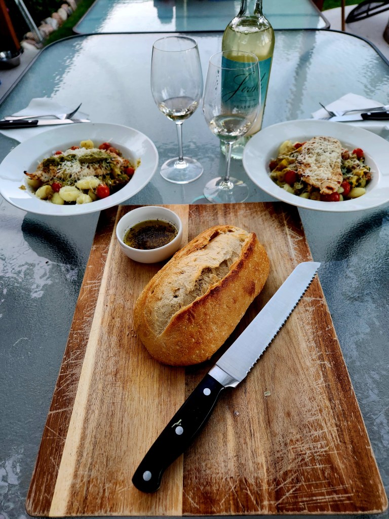  A beautiful meal setting with freshly baked bred and two bowls of Mediterranean pasta topped with cheese and two glasses of white wine.