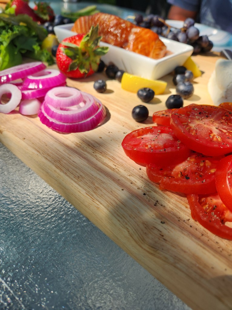 A breakfast board with tomatos, purple onion, smoked salmon, arugula, strawberries, lemon, grapes, assorted cheeses, and blueberries.